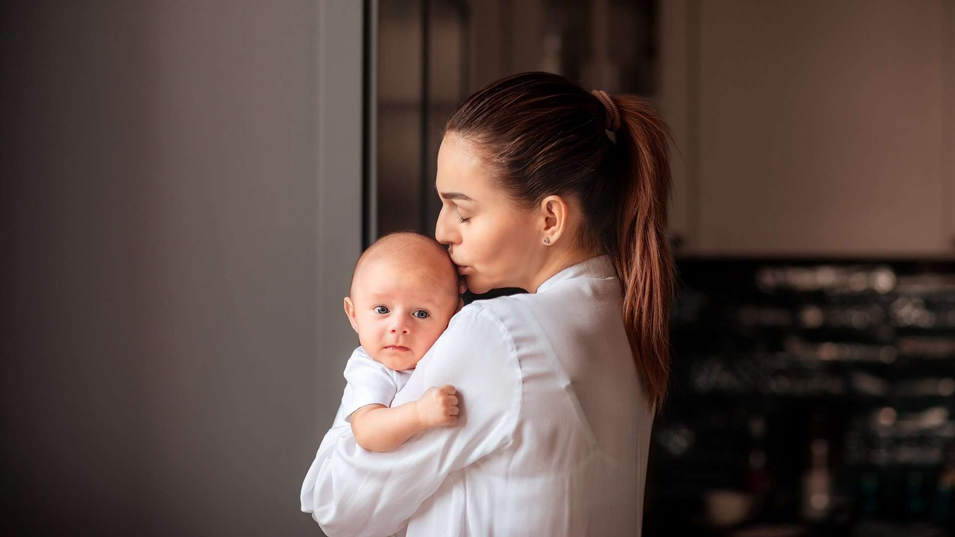 Mama caluje dziecko trzymane na rekach w glowe